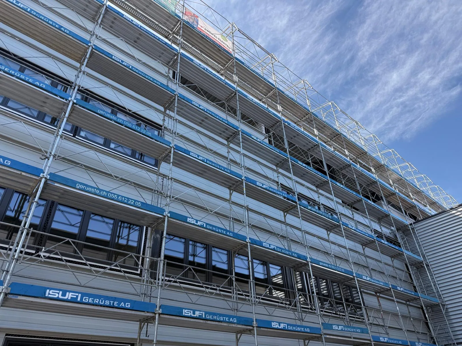 Fassadengerüst mit umlaufendem Seitenschutz Fassadengerüst mit umlaufendem Seitenschutz. Die eingerüstete Fassade zeigt Arbeitsbeläge und Geländer für den Zugang zu verschiedenen Gebäudehöhen. Referenzfoto der Isufi Gerüste AG.